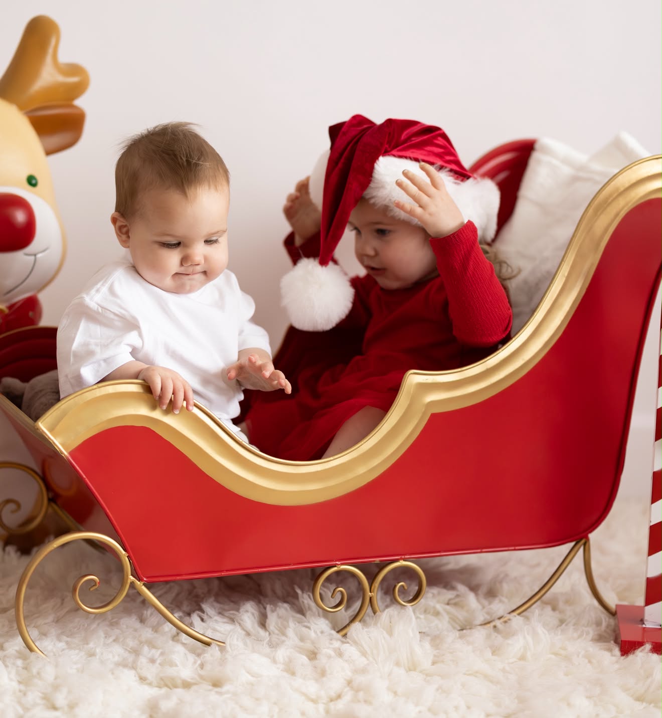 Toddler in a red velvet Santa hat holding onto her hat while sitting in the Christmas sleigh