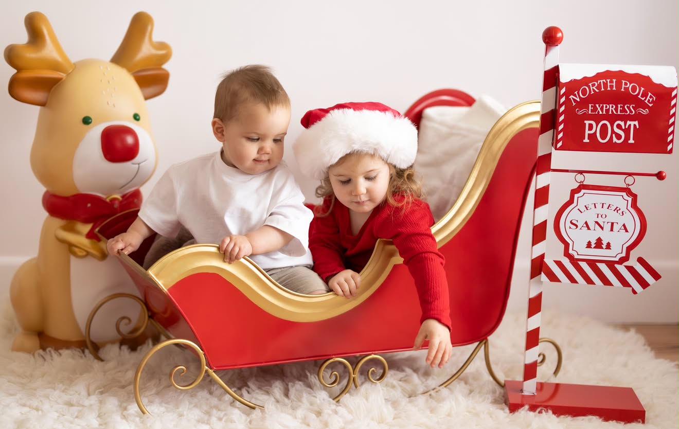 Siblings in a red Christmas sleigh beside a North Pole post box